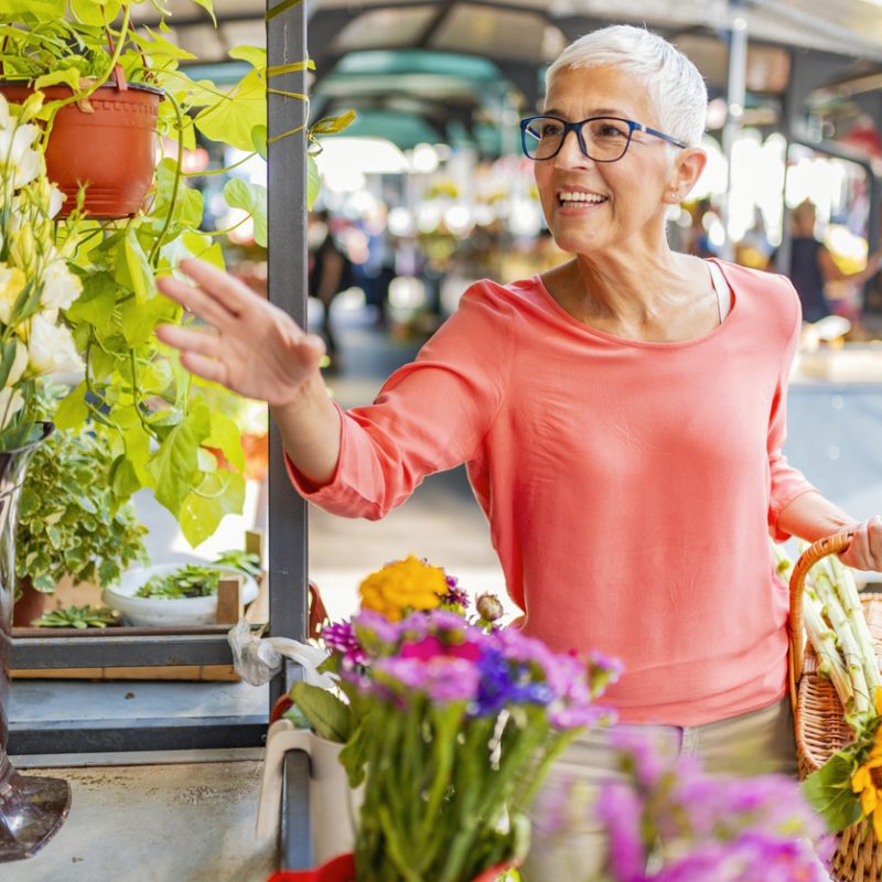 A smiling older woman reaches for a beautiful hanging plant at a stall in a farmer’s market.