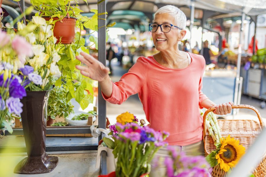 A smiling older woman reaches for a beautiful hanging plant at a stall in a farmer’s market.