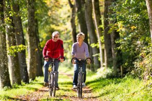 Senior man and woman exercising with bicycles outdoors, they are a couple smiling in the sunshine as they bike along a wooded trail.
