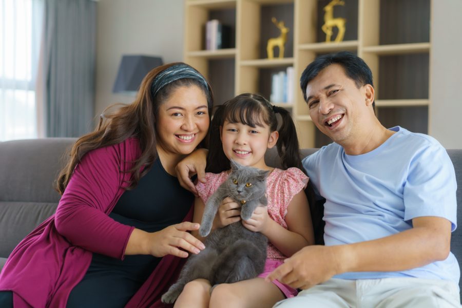A happy family sits together on a couch in their apartment, with the daughter holding a cat.